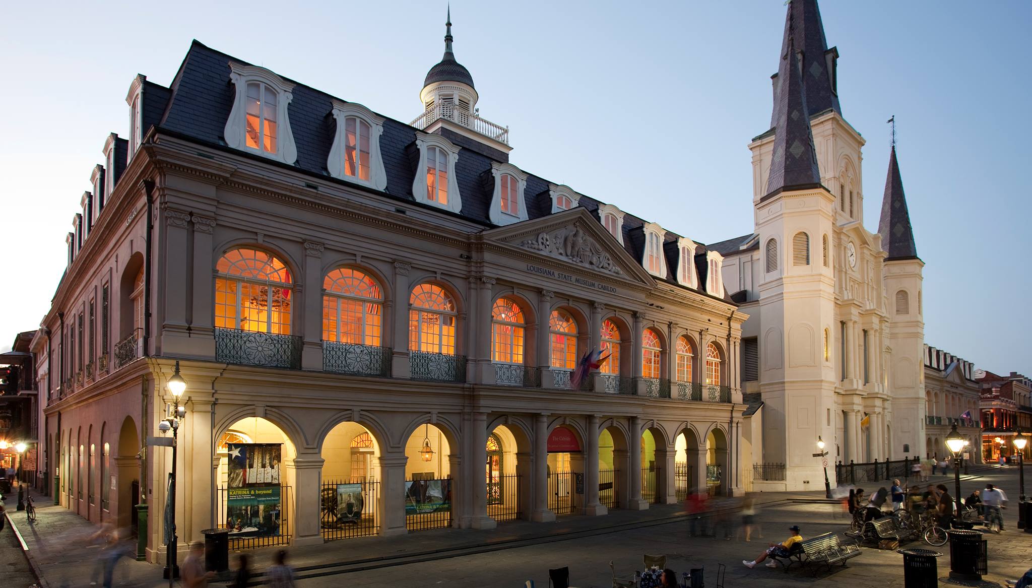 A large, historic building with arches and a balcony, situated on a street with a church steeple in the background.
